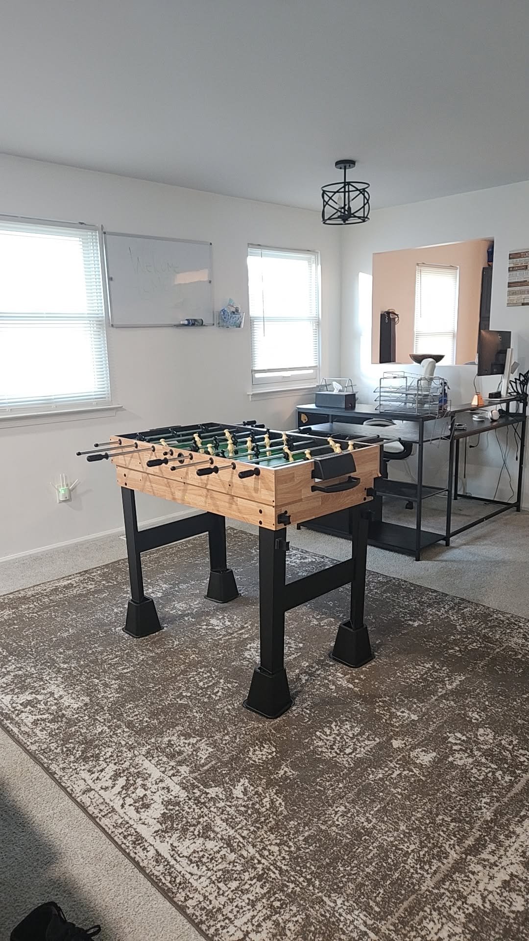 Foosball table with wooden frame and black legs positioned on a patterned area rug in a game room with desk and windows