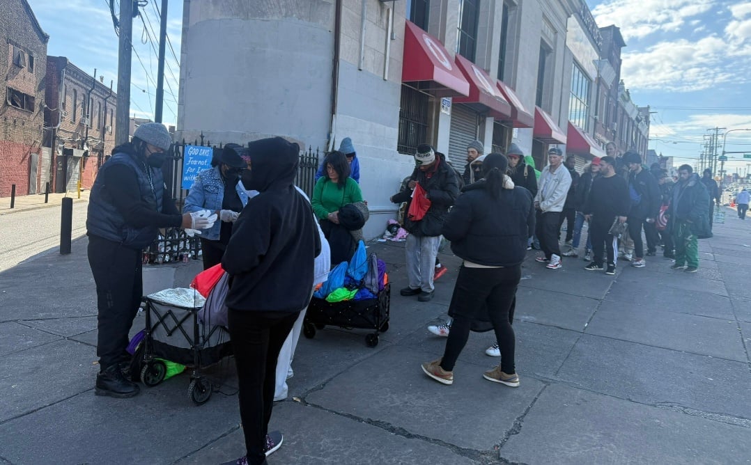 Large crowd of people waiting in line on a urban street during daytime with buildings and storefronts in background